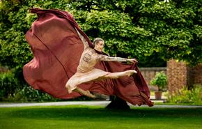 A female dancer with blonde hair and wearing a brown dress and cloak leaps in the air in a beautiful garden.